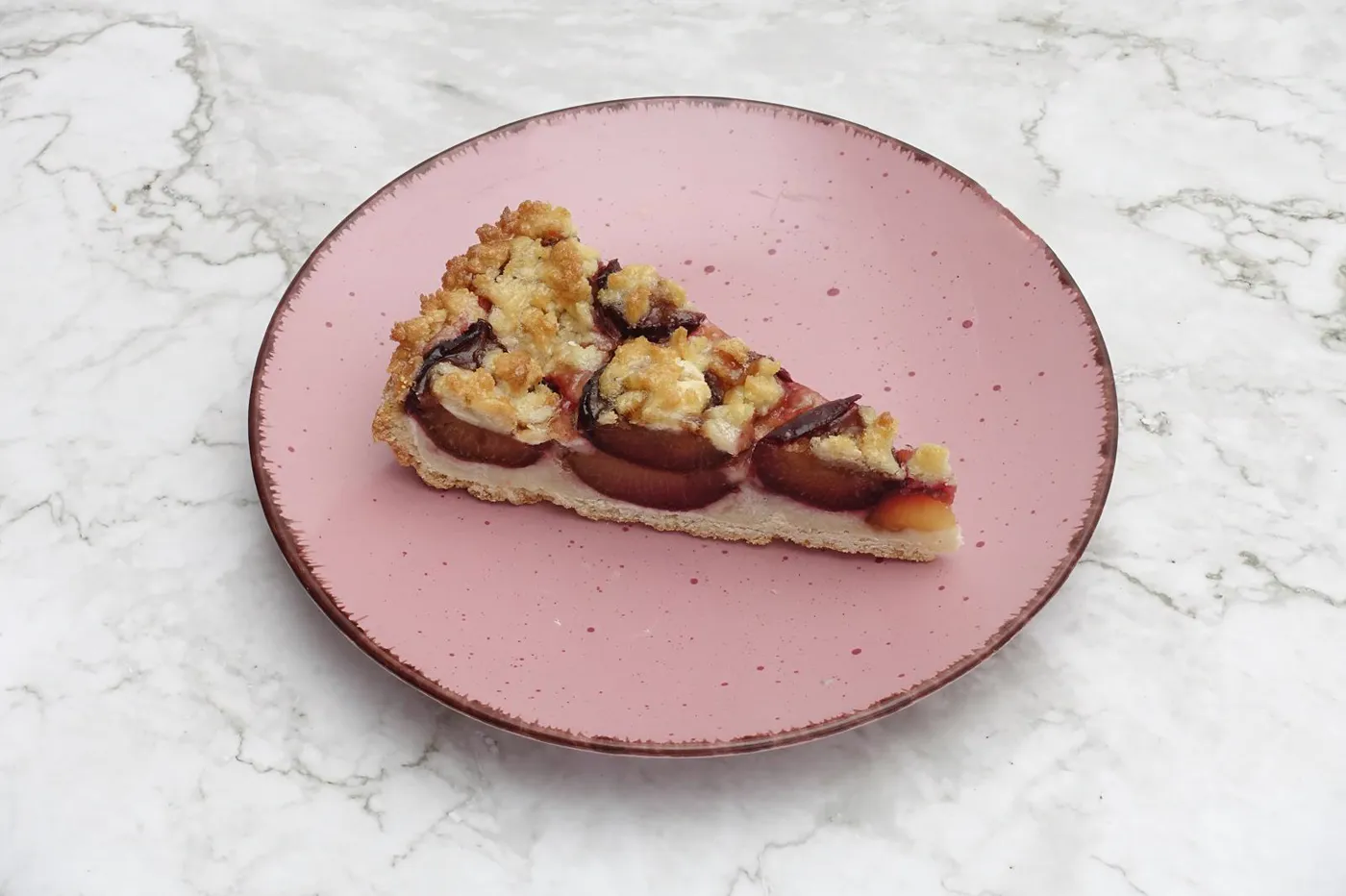 Ein Stück Pflaumen-Streuselkuchen auf einem rosa Teller auf Marmor
