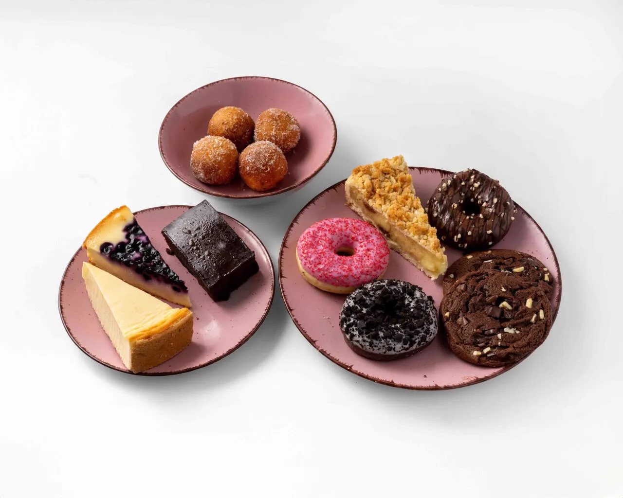 Zwei rosa Teller mit Cheesecakes, Brownie, Donuts, Apfelkuchen, Cookie und einer Schale Quarkbällchen